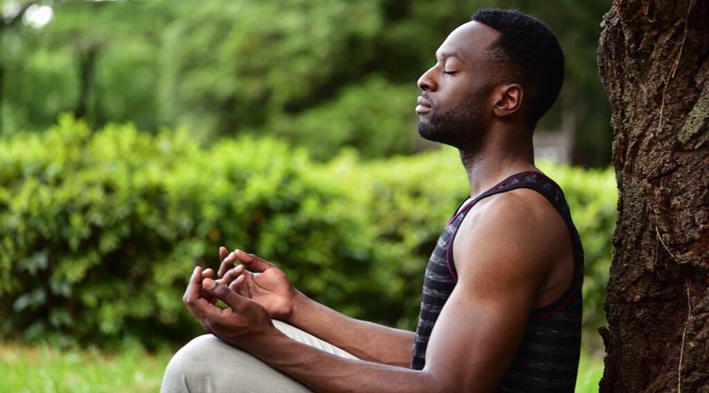 Black man sitting and meditating by a tree in what appears to be a garden.