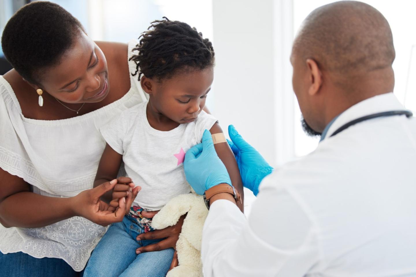 Black doctor giving out a vaccination to a black child sitting on her mother's thigh
