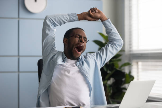 Tired african American male worker yawn stretch in office chair overwhelmed with work at workplace, exhausted biracial young man employee sign feel fatigue after long day, having sleep deprivation